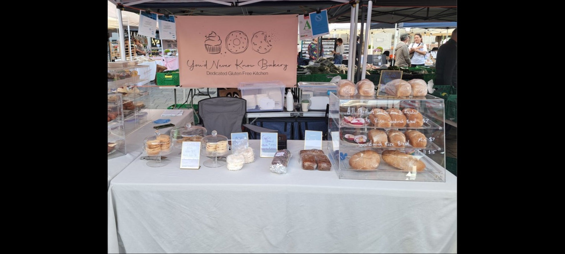 Gluten free bakery stand with various baked goods under a striped umbrella at an outdoor market.